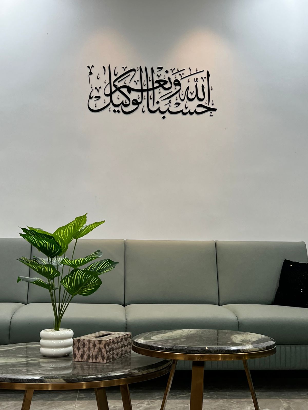 Modern Arabic calligraphy wall art in matte black acrylic displaying "Hasbunallahu Wa Ni’mal Wakeel" above a stylish sofa in a contemporary living room.